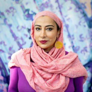 Najeeba Syeed is wearing a pink heaadscarf, round gold earrings, and a purple shirt while posing against a blue and purple background.